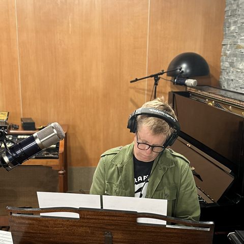 Jeff Babko at the studio keyboards in headphones and an olive jacket, reading from a score.