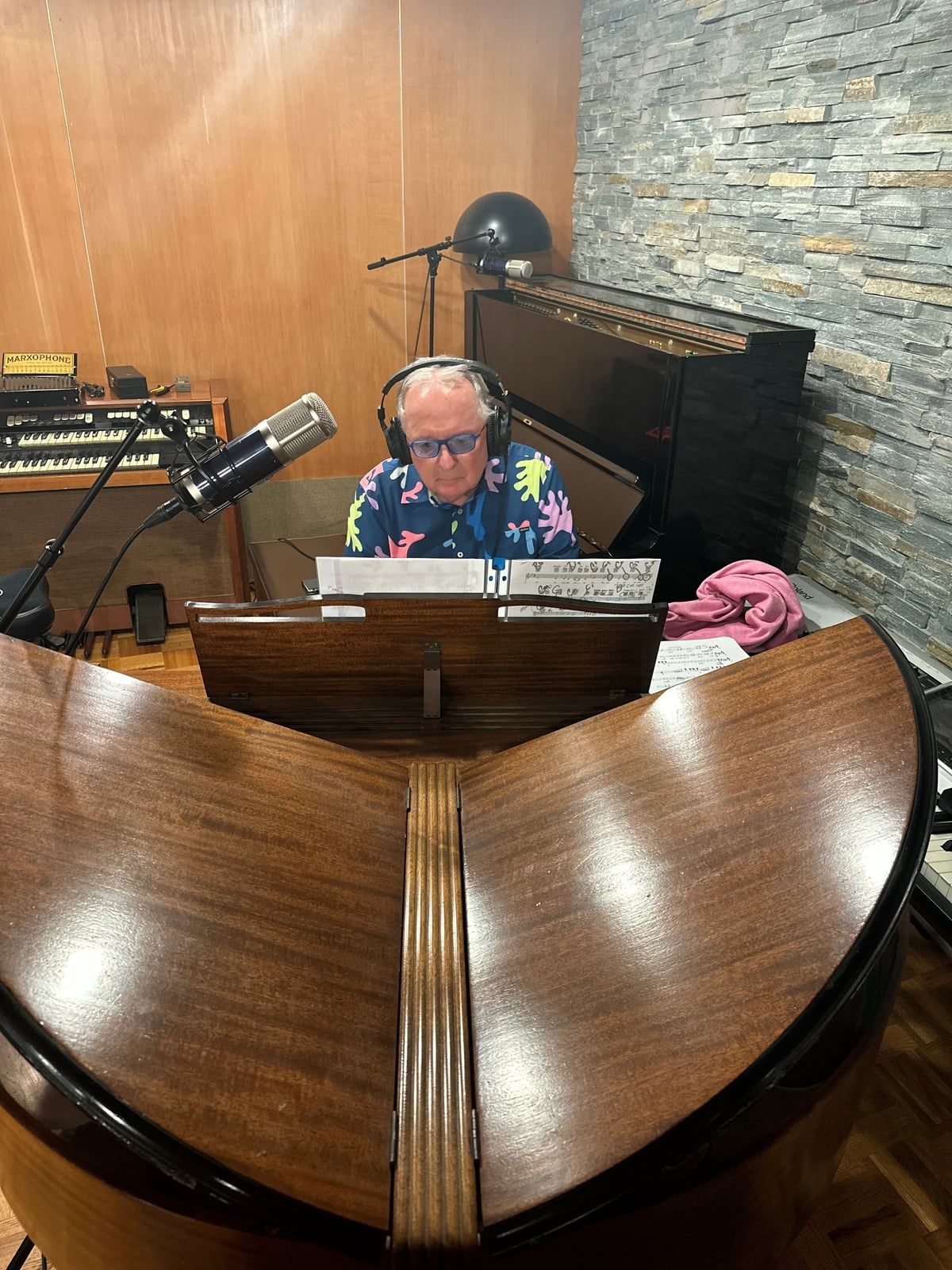 Dr. Dennis Michels at a grand piano in the studio, wearing headphones and a bright patterned shirt, the open piano lid rising like wings in the foreground.