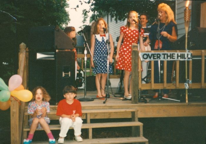 The young Michels sisters singing into microphones on an outdoor wooden stage at a community event, two smaller children sitting on the steps below and balloons tied to the rail.
