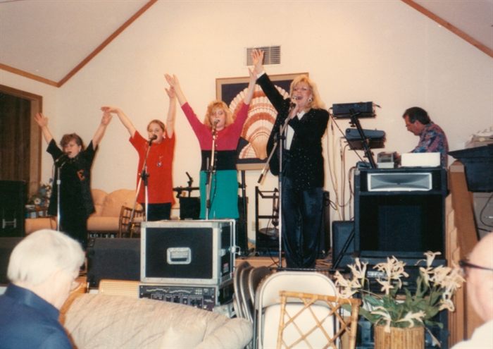 The Michels family performing in a living room with arms raised on the last note, Doc at a keyboard to the side and an audience seated on the sofa.