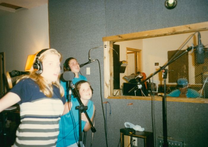 Three of the Michels sisters as girls, in headphones at microphones in a recording studio, the control room window behind them.