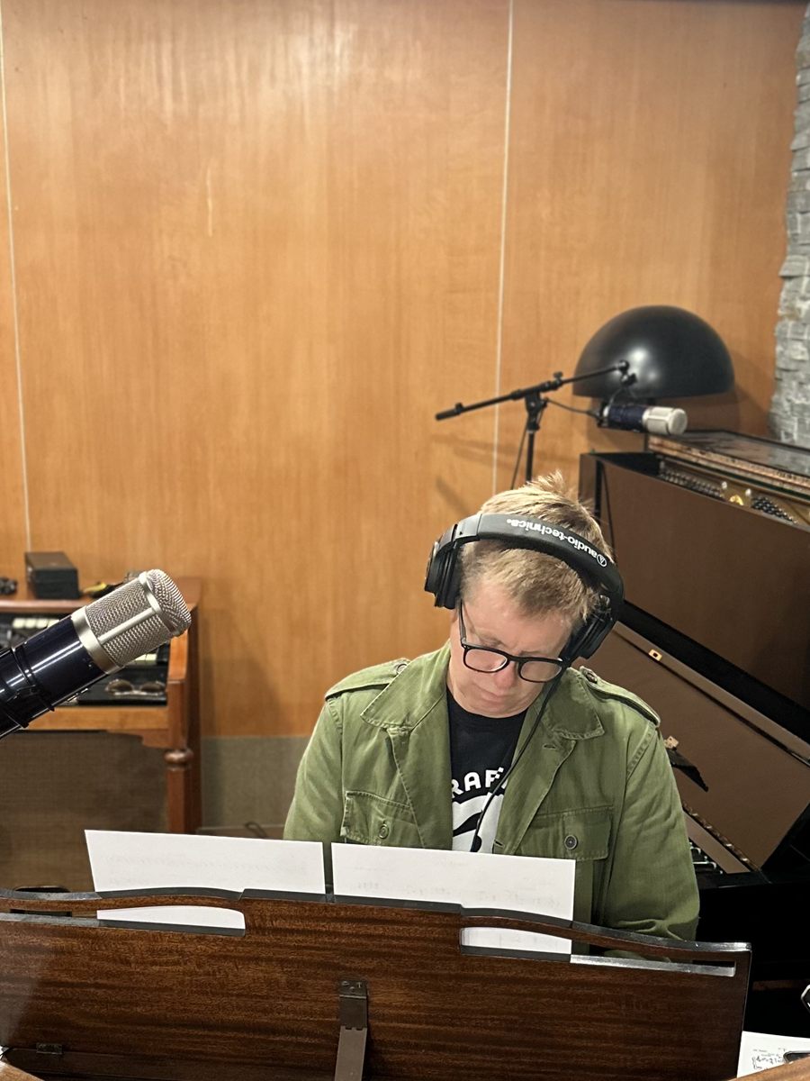 Jeff Babko at the studio keyboards in headphones and an olive jacket, reading from a score on the stand.