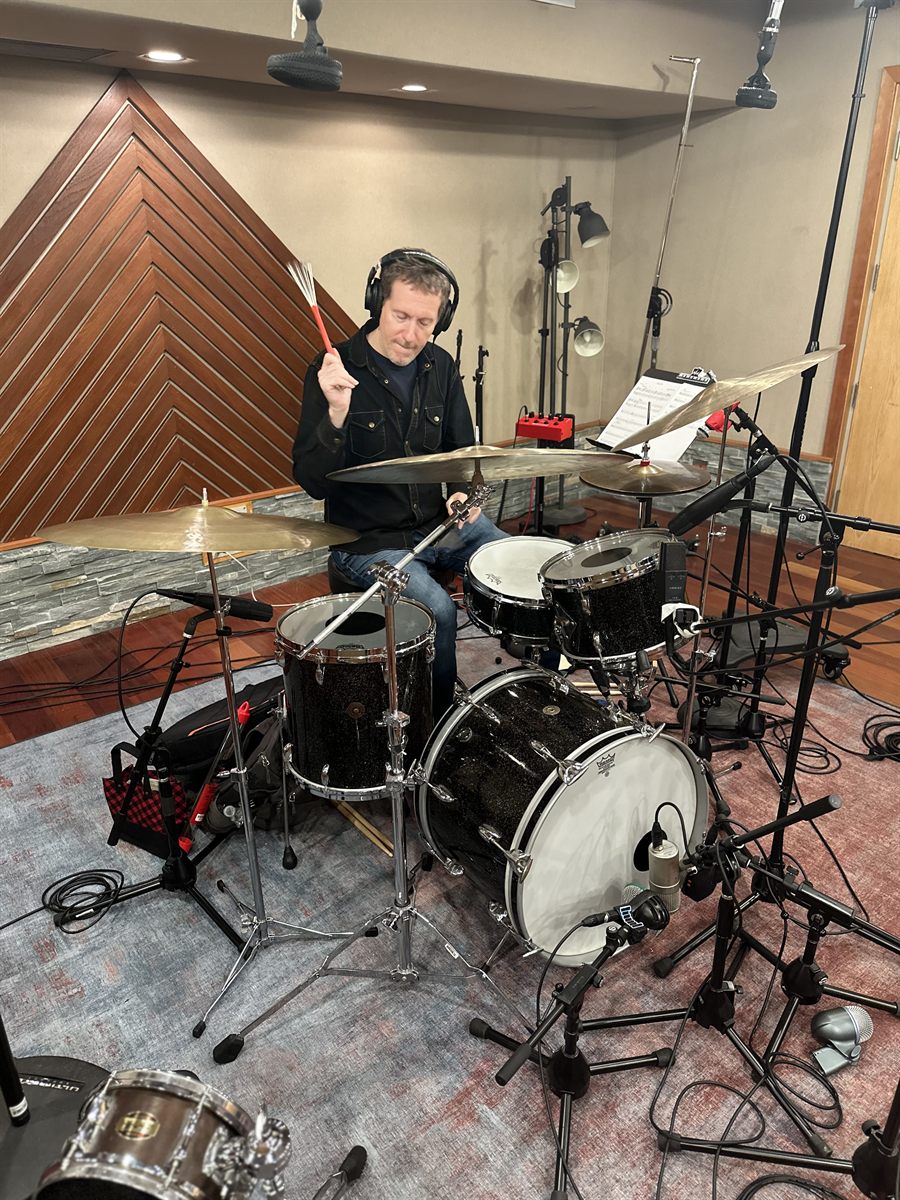 Zach Danziger behind his drum kit in the studio, brushes raised, headphones on, a marked-up chart on the stand beside him.