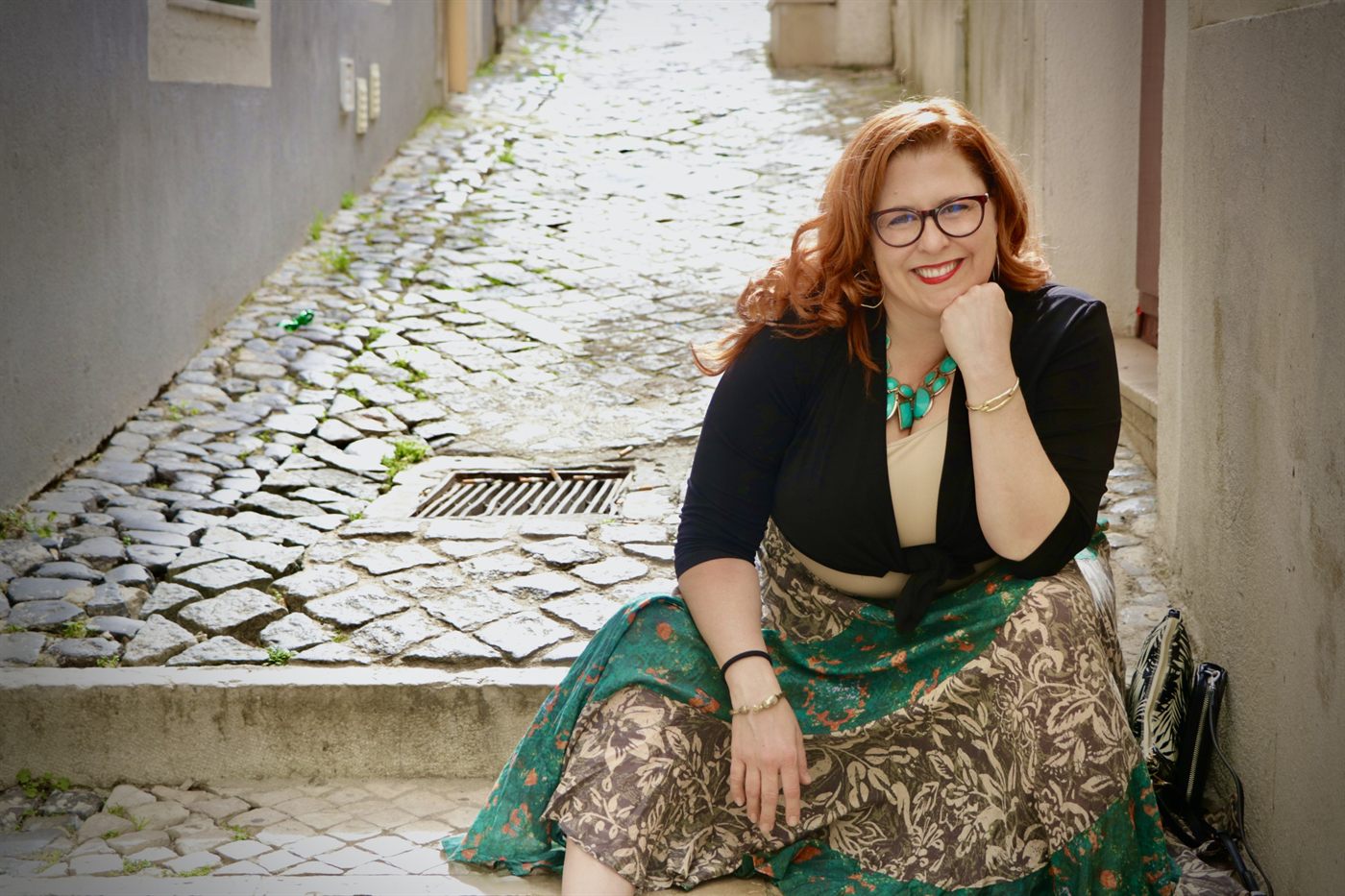 Jenn Quader sitting on a step in a narrow cobbled alley, chin on her hand, in a green and cream patterned skirt.