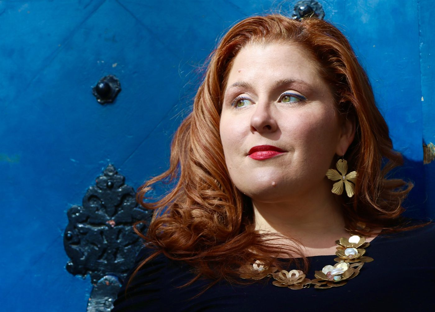 Jenn Quader in profile against a blue studded door, looking upward, wearing a gold flower earring and a gold and pearl necklace.