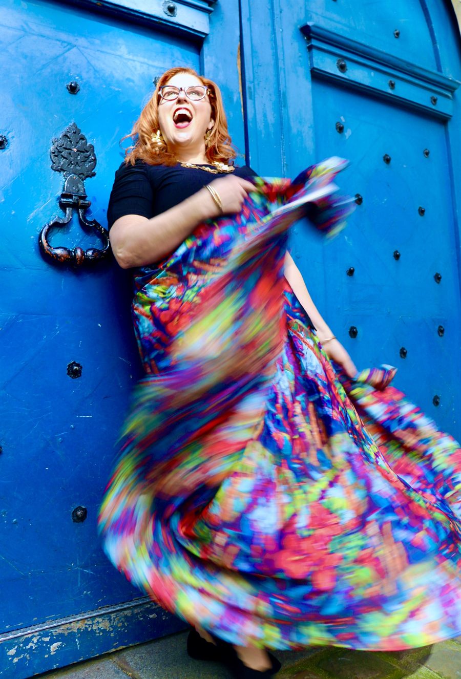 Jenn Quader laughing with her head thrown back, spinning so that her brightly patterned skirt flares out around her, against the blue studded door from the album cover.