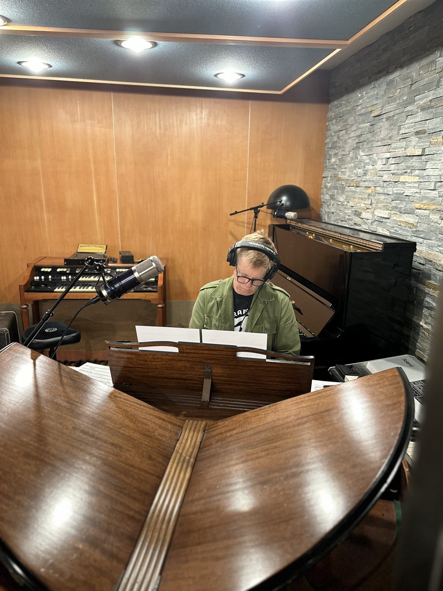 Jeff Babko at the studio grand piano in headphones and an olive jacket, reading from a score, the open butterfly music desk of the piano filling the foreground.