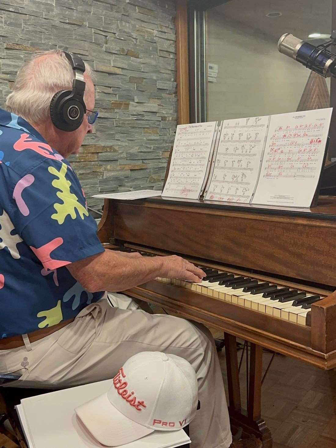 Dr. Dennis Michels at the studio upright in headphones and a bright patterned shirt, marked-up sheet music on the stand.